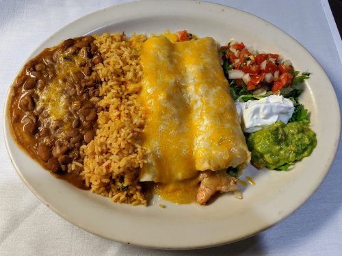 A plate of enchiladas topped with cheese, served with rice, beans, guacamole, salsa, and sour cream.