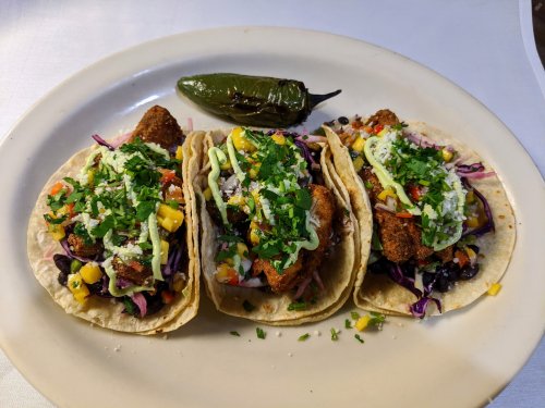 Three tacos filled with fish, mango, and cabbage, topped with green sauce and cilantro, served with a jalapeño on the side.