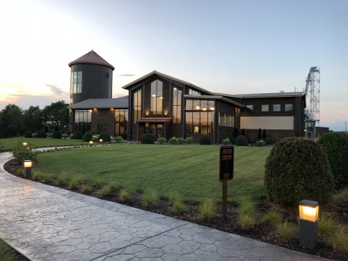 A modern visitor center with large windows, surrounded by landscaped gardens and a pathway, under a sunset sky.