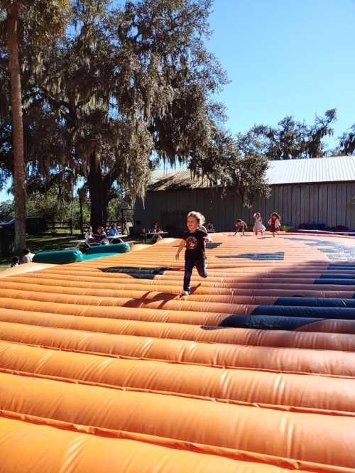 A child runs joyfully on a large inflatable bounce pad under a clear blue sky, surrounded by trees and other kids.