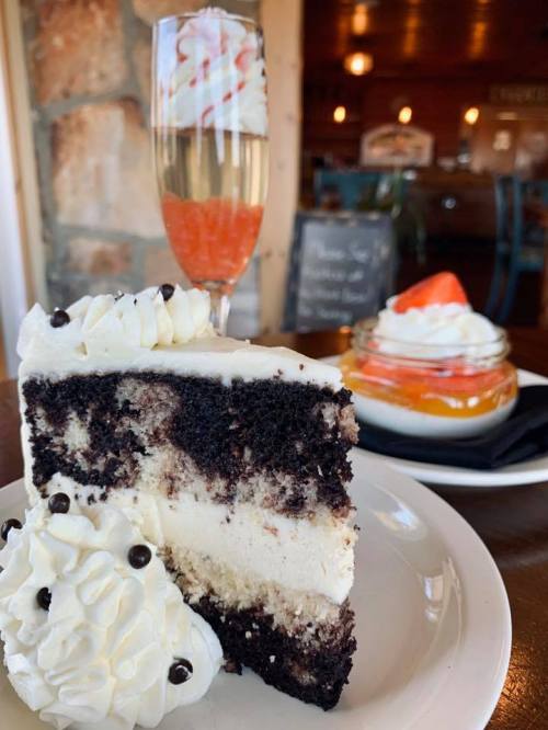 A slice of layered chocolate cake with cream, whipped topping, and a dessert glass with fruit and a drink in the background.