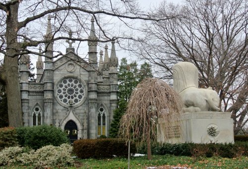 A gothic-style building with a large rose window, next to a stone lion statue and bare trees in a park setting.