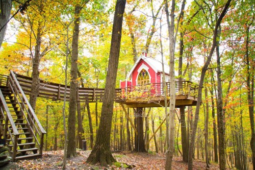 A whimsical red treehouse nestled among vibrant autumn trees with colorful leaves and a wooden walkway.