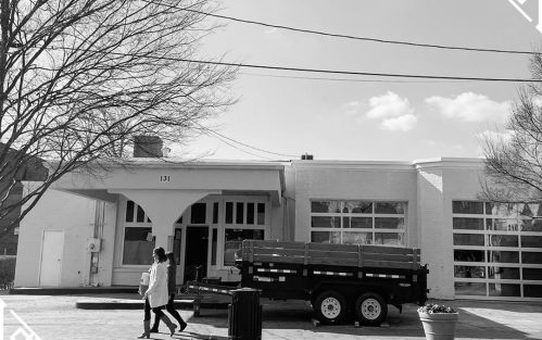 A person walks past a white building with large windows and a truck parked outside, under a clear sky.