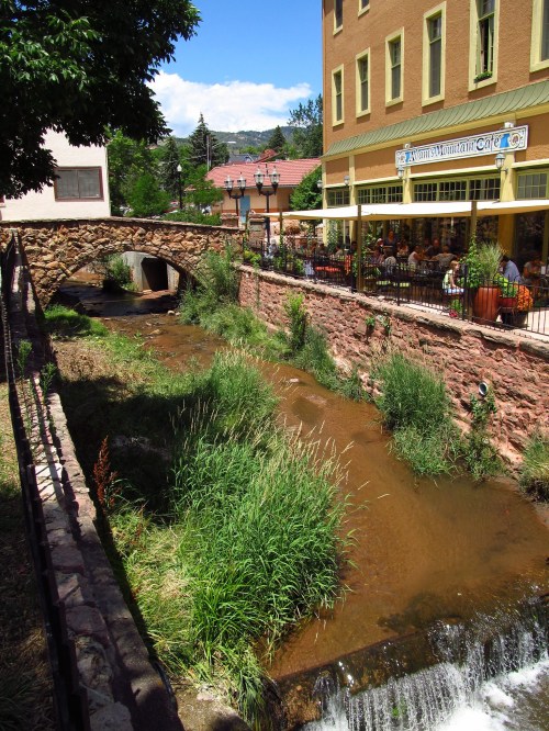 A quaint stone bridge over a stream, with a café and greenery in the background on a sunny day.