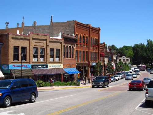 A sunny street scene featuring historic buildings, shops, and parked cars in a small town.