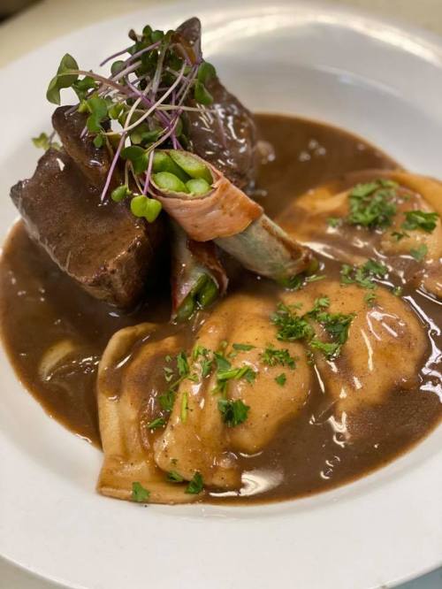 A plate of tender beef with green beans wrapped in bacon, served with dumplings in a rich brown sauce and garnished with herbs.