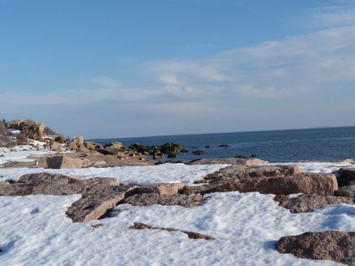 Rocky shoreline with snow, calm blue water, and a clear sky in the background.