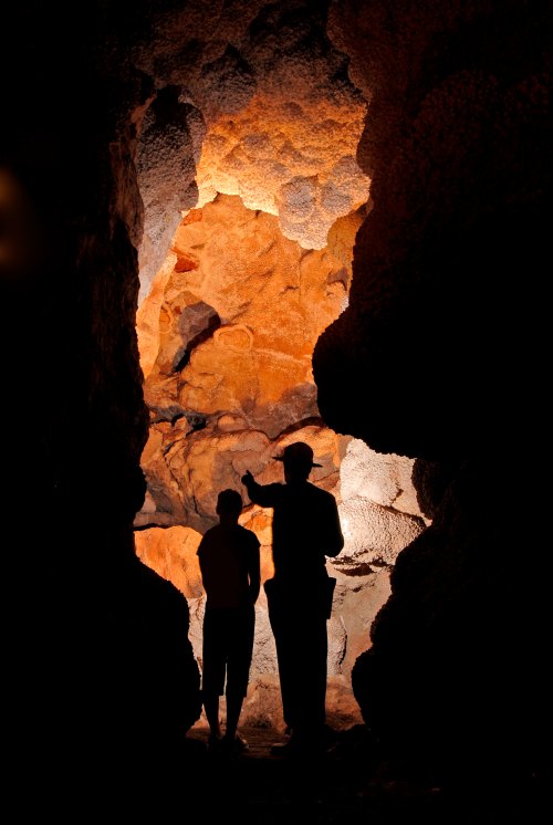 Two silhouetted figures stand in a cave, illuminated by warm light reflecting off the rocky walls.