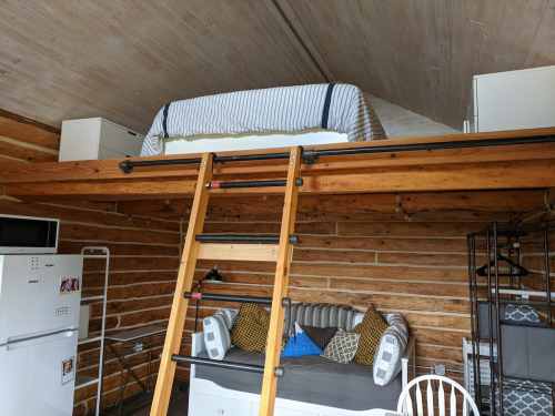 A cozy lofted bed above a seating area in a wooden cabin, with a ladder for access and a small kitchen in the background.