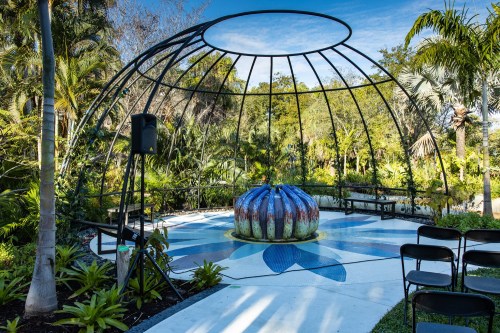 A circular garden pavilion with a colorful fountain surrounded by lush greenery and empty chairs.