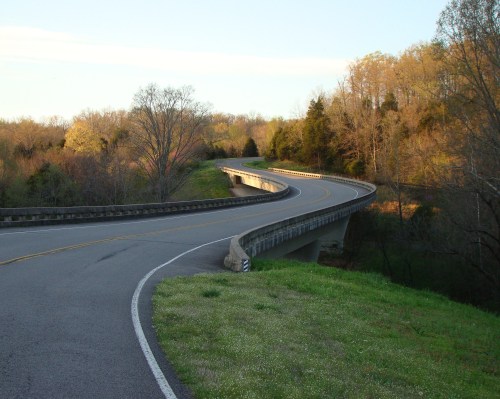 A winding road curves over a bridge, surrounded by trees and greenery under a clear sky.