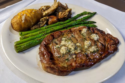 A plate featuring a grilled steak with herb butter, asparagus, baked potato, and assorted mushrooms.