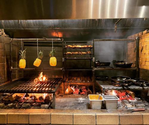 A vibrant kitchen with a grill, hanging pineapples, and various dishes cooking over an open flame.