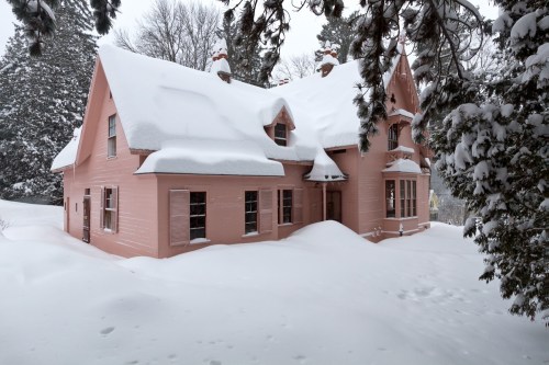 A pink house covered in snow, surrounded by tall trees, with snowdrifts nearly reaching the windows.
