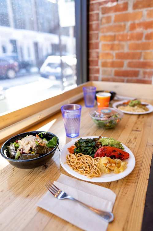 A wooden table with two plates of colorful food, a bowl of salad, and glasses of water, near a window with a view.