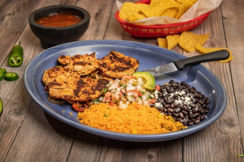 A blue plate with grilled chicken, rice, black beans, pico de gallo, and a knife, with chips and salsa in the background.