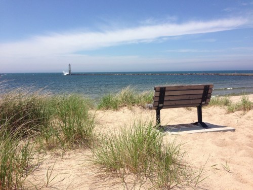 A wooden bench on sandy beach grass, overlooking a calm sea and distant lighthouse under a clear blue sky.