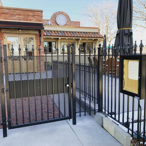 Black metal gate leading to a brick building with a sign, surrounded by a patio area and trees in the background.