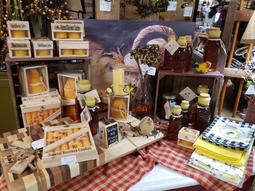 A display of beeswax candles, honey jars, and decorative items on a checkered tablecloth in a rustic setting.