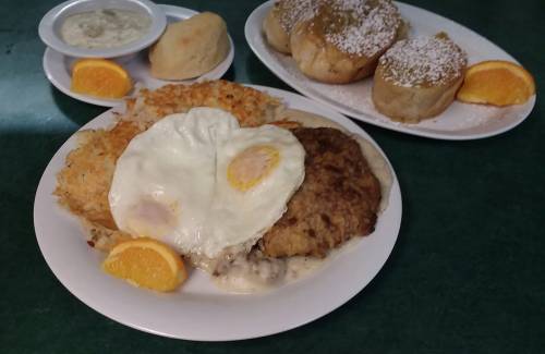 A plate of chicken-fried steak with eggs, hash browns, and orange slices, alongside cinnamon rolls and dipping sauce.