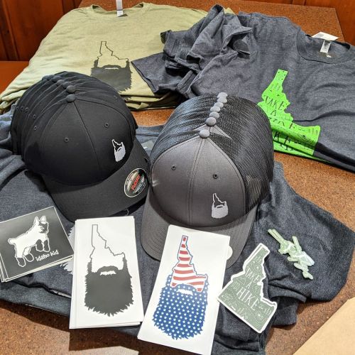 A collection of hats and t-shirts featuring Idaho-themed designs, along with stickers and decals on a wooden table.