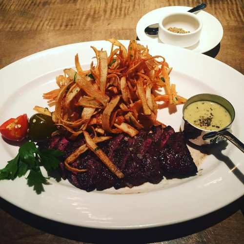 A plate of grilled steak with crispy fries, a side of sauce, and garnishes of tomato and herbs.