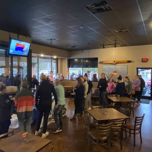 A busy restaurant interior with a crowd gathered around a table, chatting and waiting in line.