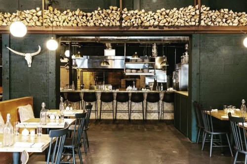 A modern restaurant interior featuring wooden decor, a visible kitchen, and tables set for dining.