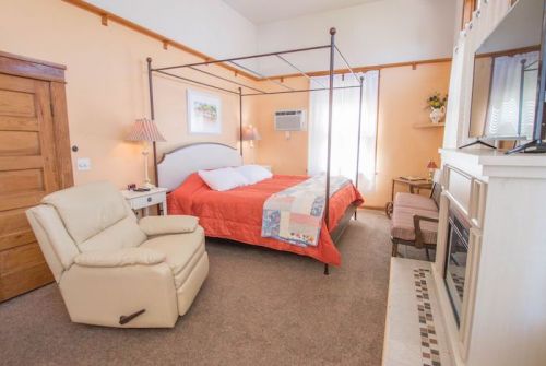 Cozy bedroom featuring a four-poster bed, armchair, and fireplace, with warm orange walls and natural light.