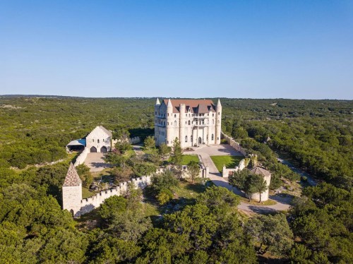 Aerial view of a large, castle-like structure surrounded by greenery and trees under a clear blue sky.
