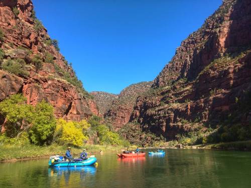 Two colorful rafts float on a calm river surrounded by steep, red rock cliffs and lush greenery under a clear blue sky.