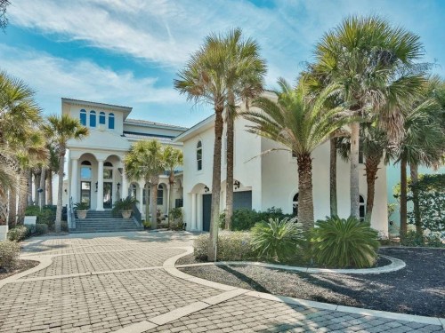 A modern white house surrounded by palm trees and a paved driveway under a clear blue sky.
