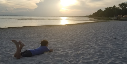 A child lies on the sandy beach, watching the sunset over the water, with trees and a pier in the background.
