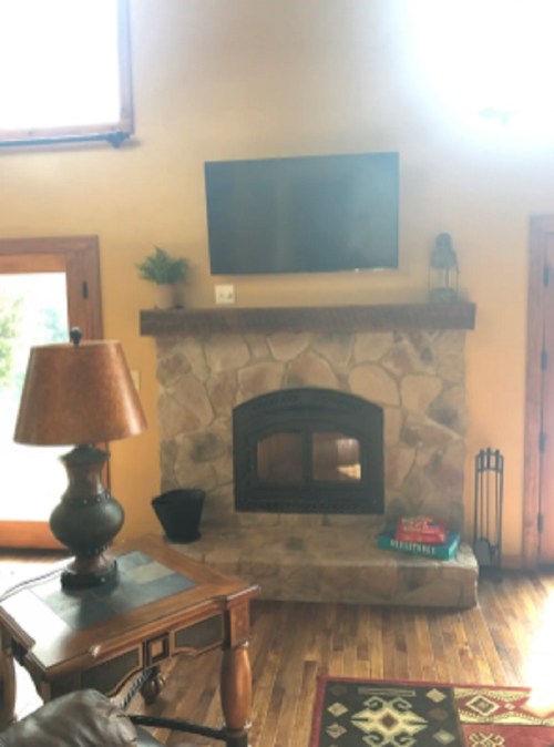 Cozy living room with a stone fireplace, mounted TV, wooden furniture, and a lamp, featuring warm, inviting decor.