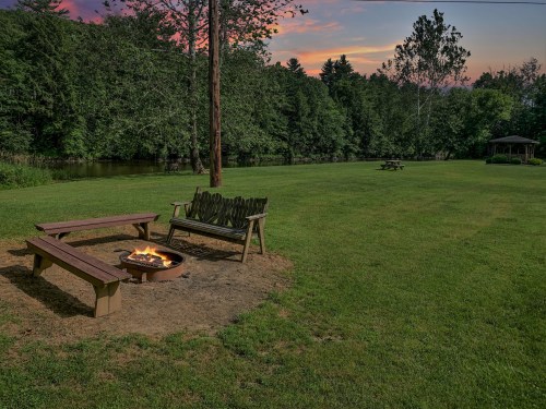 A serene outdoor scene featuring a fire pit, wooden benches, and lush greenery under a colorful sunset sky.