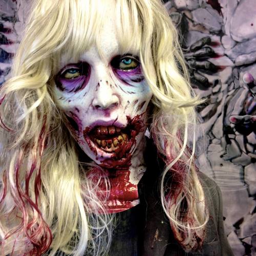 A creepy zombie figure with pale skin, long blonde hair, and bloodied features, set against a dark, textured background.