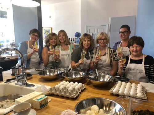 A group of seven women in aprons hold drinks, smiling in a kitchen with bowls and ingredients on the counter.