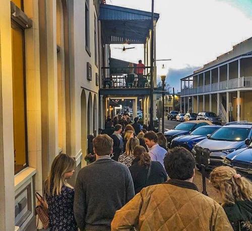 A long line of people waiting outside a building, with cars parked along the street and a sunset in the background.