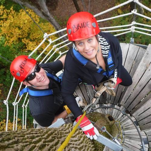 Two people in helmets, one labeled "BRIDE," climbing a tree platform with ropes and harnesses in a forested area.