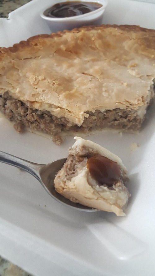 A slice of pie with a flaky crust, revealing a meat filling, next to a spoonful of the filling and a small cup of sauce.
