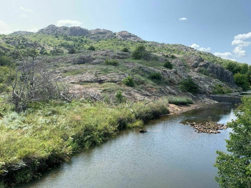 A serene river flows through a rocky landscape, surrounded by lush greenery and distant hills under a clear blue sky.