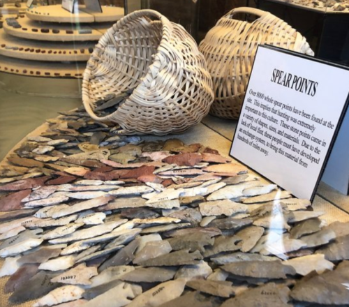 Display of various spear points and woven baskets in a glass case, with an informational sign about their history.