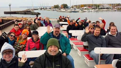 A group of smiling people on a boat, waving at the camera, with a marina and boats in the background.