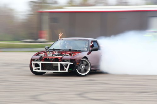 A modified car drifts on a track, emitting smoke, with a driver giving a peace sign out the window.