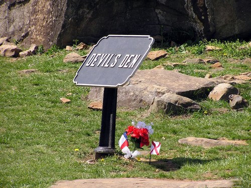 Sign for "Devil's Den" with flowers and flags nearby, set against a rocky landscape.
