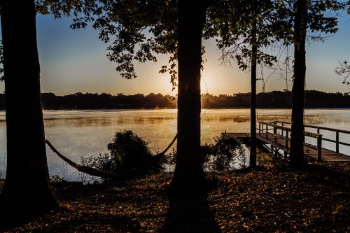 Sunrise over a calm lake, with a hammock between trees and a wooden dock in the foreground.