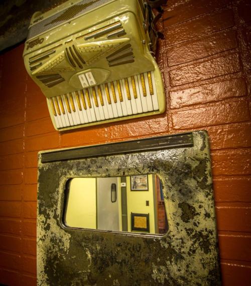 An old accordion hangs above a weathered metal panel with a small mirror, set against a brick wall.