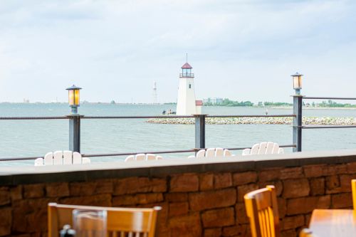 View of a lighthouse by the water, with a railing and chairs in the foreground, under a cloudy sky.