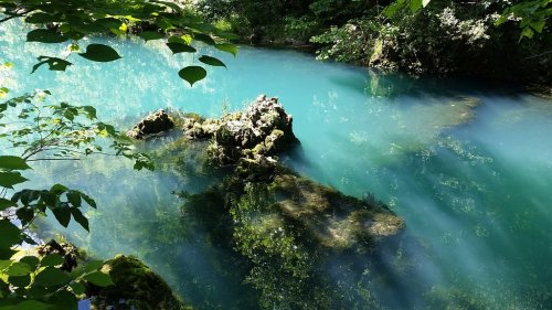 A serene turquoise river with rocky outcrops and lush greenery reflecting in the water. Sunlight filters through the trees.